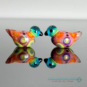 May include: Two small glass bird figurines featuring a vibrant pattern of pink, orange, yellow, and blue circular spots. Each bird has a solid blue head, a black beak, and black eyes. The text SOJKA LAMPWORK STUDIO crafted by flame is visible.
