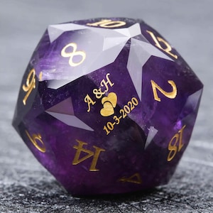 May include: A faceted, purple gemstone D20 die with gold-colored numerals. The die is engraved with the initials "A & H", two heart symbols, and the date "10-3-2020". The die is a unique item for tabletop gaming.