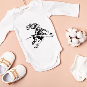 May include: White baby onesie with a black dinosaur graphic. The onesie has long sleeves and a snap closure. The image also includes white baby shoes with tan laces, a cotton flower, and a white baby sock.