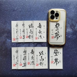 以下が含まれることがあります： 金色のフレームが付いた透明な携帯電話ケース。日本語の書道が書かれた白いカードが入っています。日本語の書道が書かれた小さな白いカードがいくつか下に配置されています。背景は濃い青色の斑点のある表面です。