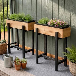 Op de afbeelding: Twee houten rechthoekige plantenbakken met zwarte metalen poten, elk gevuld met groen en bloemen. De plantenbakken zijn gemaakt van lichtgekleurd hout en staan op een grijs tapijt. Een gieter en kleine potplanten staan op de voorgrond.