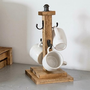 May include: A wooden mug tree with a dark metal top and hooks, holding four white ceramic mugs. The tree has a square base and a rustic, farmhouse style. The mugs have a speckled design.
