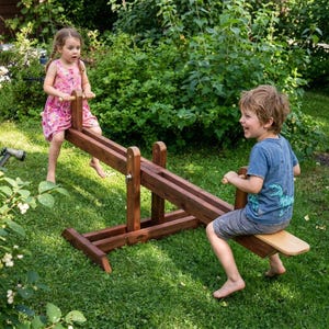 Könnte beinhalten: Eine Holzwippe in einem grünen Garten. Zwei Kinder spielen auf der Wippe, die aus dunkelbraunem Holz gefertigt ist. Die Kinder lächeln und scheinen Spaß an dem Spielgerät im Freien zu haben.