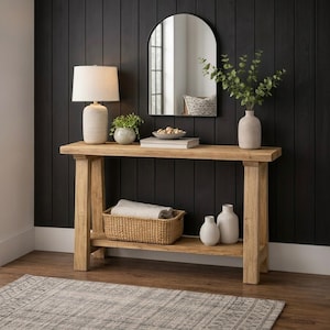 Op de afbeelding: Een houten console met een lichte afwerking, voorzien van een onderste plank en een rechthoekige bovenkant. De tafel is versierd met een lamp, spiegel, vazen en een gevlochten mand. De donkere muur erachter contrasteert.