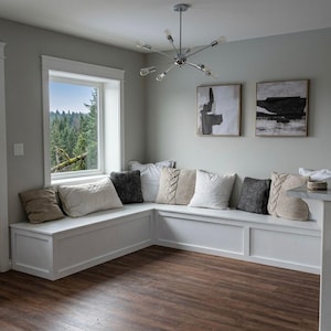 May include: A built-in white bench seat with several decorative pillows in various colors and textures. The pillows include beige, white, and gray tones. The bench is positioned beneath a window with a view of a forest.