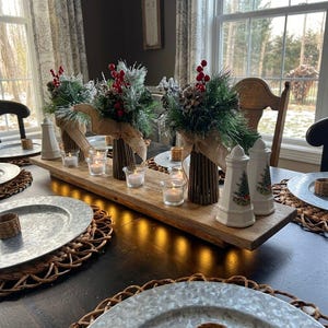 Puede incluir: Centro de mesa rústico de madera con tres arreglos florales decorativos en jarrones de ramitas con cinta de arpillera. Pequeñas velas votivas y dos saleros y pimenteros con forma de faro también están en la bandeja de madera. La mesa está puesta para una comida festiva.