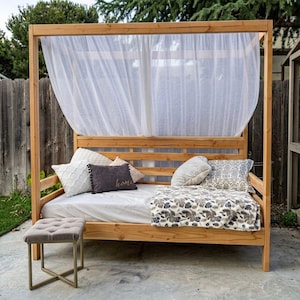 May include: A wooden outdoor daybed with a canopy, featuring white sheer curtains. The bed is furnished with pillows and a floral patterned blanket. A small, tufted ottoman sits in front.