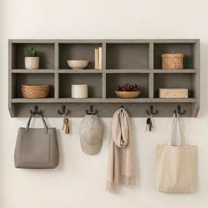 May include: A gray wooden wall shelf with eight cubbies, holding decorative items like a potted plant, bowls, books, and a woven basket. Below, six black hooks hold a tote bag, keys, a hat, and a scarf. A neutral, organized entryway.