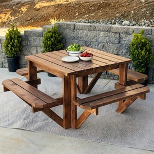 May include: A square wooden picnic table with attached benches. The table and benches are made of stained wood and are arranged around the table. A plate, bowl of salad, and bowl of strawberries sit on the table.