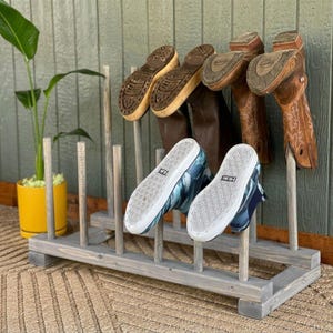 May include: A gray wooden boot rack holding various footwear, including brown cowboy boots, dark brown boots, and blue and white sneakers. The rack is rectangular with vertical supports and is placed on a woven rug.