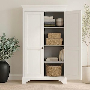 May include: A white wooden cabinet with open doors, revealing shelves with folded linens, woven baskets, and books. A black vase with green foliage and a potted plant with long leaves are on either side.