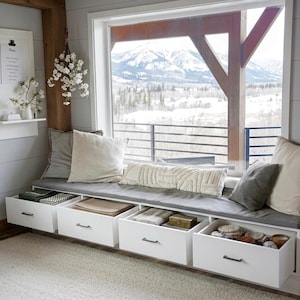 May include: A window seat with a gray cushion and several pillows. Below the seat are white drawers with black handles, some open to reveal contents. The window offers a view of snow-covered mountains.