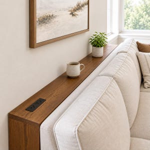 Puede incluir: Una mesa consola de madera larga y estrecha colocada detrás de un sofá beige. La mesa presenta un acabado de grano de madera oscuro, una regleta eléctrica negra integrada, una taza de cerámica y una pequeña planta verde en maceta.
