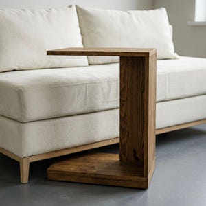 May include: A dark brown wooden side table. The table has a rectangular top, a vertical support, and a base. The table is positioned beside a cream-colored sofa. The table's dimensions are approximately 76 cm high.