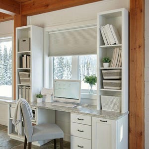 May include: A white home office desk with a computer, keyboard, and chair. The desk has built-in shelving units on either side, with storage bins, books, and decorative plants. A window with a view of a snowy landscape is behind the desk.