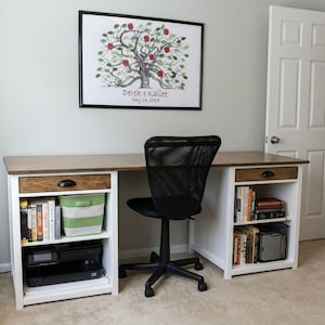 May include: A home office setup featuring a wooden desk with white storage compartments and a black mesh office chair. The desk has a framed tree art piece above it, and the storage includes books, a printer, and a striped basket.