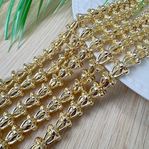 May include: Close-up of several strands of gold-coloured decorative trim. The trim has a repeating floral or leaf-like design. The strands are arranged diagonally, with a white plate in the background. Ideal for craft projects.