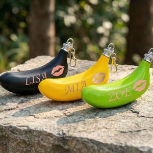 Könnte beinhalten: Drei bananenförmige Schlüsselanhänger in Schwarz, Gelb und Grün, jeweils mit silberner Kappe und goldfarbenem Verschluss. Der schwarze Schlüsselanhänger hat den Namen "LISA" und einen rosa Lippenabdruck. Der gelbe Schlüsselanhänger hat "MIA" und einen Lippenabdruck. Der grüne Schlüsselanhänger hat "ZOE" und einen Lippenabdruck.