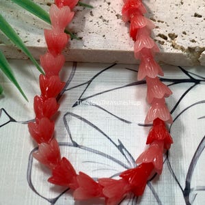 May include: A strand of beads shaped like small tulip flowers in variegated shades of red and pink. The beads transition from deep red to lighter coral and pale pink hues. The text Variegated Red and TimelessTreasuresHup are visible.