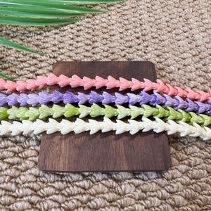 May include: Four strands of small, flower-shaped beads arranged horizontally. The strands are colored in light pink, lavender, light green, and cream. Each bead features a delicate, multi-petaled floral design linked together in a continuous row.