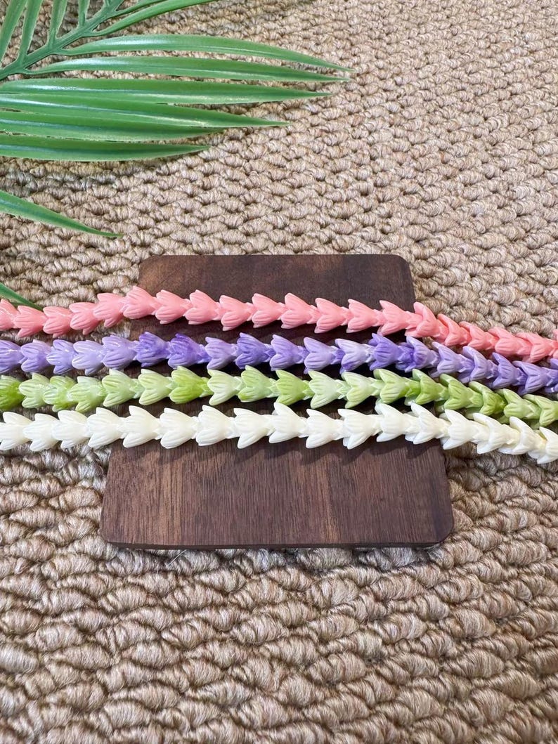 May include: Four strands of small, flower-shaped beads arranged horizontally. The strands are colored in light pink, lavender, light green, and cream. Each bead features a delicate, multi-petaled floral design linked together in a continuous row.