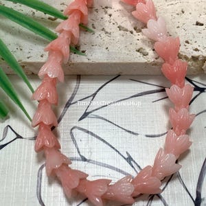 May include: A strand of pink, carved flower-shaped beads arranged in a curved line. The beads feature a translucent, glossy finish with detailed petal textures. The text "Pink" and "TimelessTreasuresHup" are visible on the image surface.