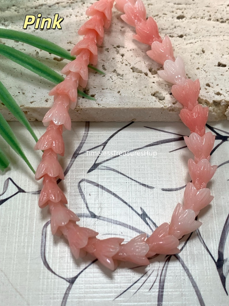 May include: A strand of pink, carved flower-shaped beads arranged in a curved line. The beads feature a translucent, glossy finish with detailed petal textures. The text "Pink" and "TimelessTreasuresHup" are visible on the image surface.