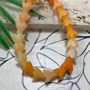 May include: A bracelet composed of translucent, flower-shaped beads in varying shades of orange, ranging from pale peach to deep amber. The beads are strung together in a circular pattern. The text "Orange" and "TimelesstreasuresHup" are visible.