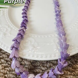 May include: A necklace composed of numerous small, carved purple flower-shaped beads strung together in a continuous strand. The beads feature varying shades of light and dark purple, creating a textured, floral appearance throughout the length of the piece.