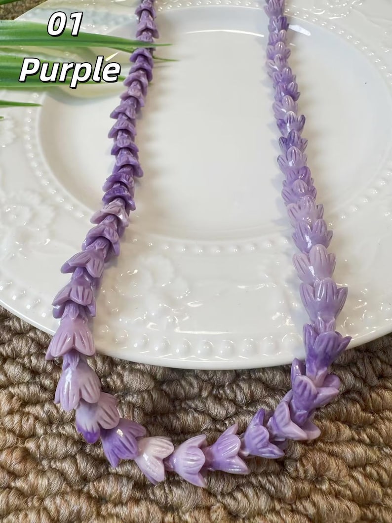 May include: A necklace composed of numerous small, carved purple flower-shaped beads strung together in a continuous strand. The beads feature varying shades of light and dark purple, creating a textured, floral appearance throughout the length of the piece.