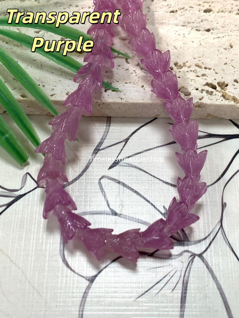 May include: A strand of transparent purple beads shaped like small flower buds. The beads are arranged in a curved line. The text Transparent Purple is visible in the upper left corner, and the text TimelessTreasuresHub is visible in the center.