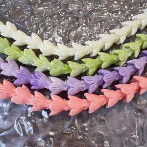 May include: Four strands of flower-shaped beads arranged in parallel rows. The top strand is cream, the second is light green, the third is lavender, and the bottom strand is coral pink. Each bead features a carved petal design in a matte finish.