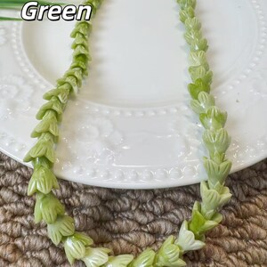 May include: A necklace composed of light green, carved floral-shaped beads arranged in a continuous strand. The text 02 Green is visible in the upper left corner. The beads feature a smooth, polished finish and are shaped like small, bell-like flowers.