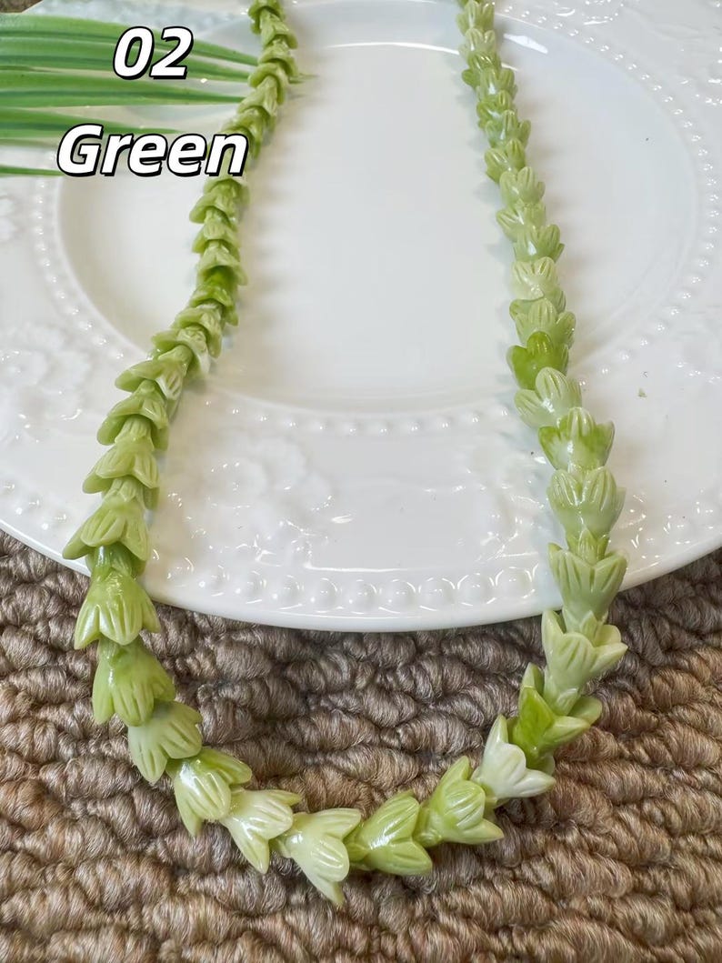 May include: A necklace composed of light green, carved floral-shaped beads arranged in a continuous strand. The text 02 Green is visible in the upper left corner. The beads feature a smooth, polished finish and are shaped like small, bell-like flowers.