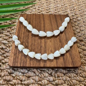 Könnte beinhalten: Zwei Stränge weißer, blütenblattförmiger Perlen, die auf einer kleinen, quadratischen Holzplatte angeordnet sind. Die Perlen sind zu einer Halskette oder einem Armband aufgereiht. Die Holzplatte hat eine satte, dunkelbraune Farbe.