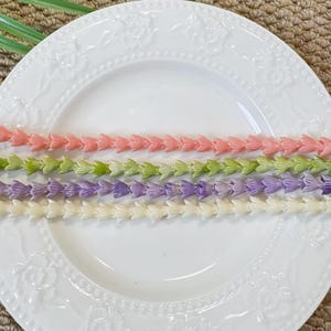 May include: Four strands of small, molded plastic flower beads arranged horizontally on a white ceramic plate with an embossed floral rim. The strands are colored in soft shades of peach, light green, lavender, and cream, each featuring a repeating petal design.