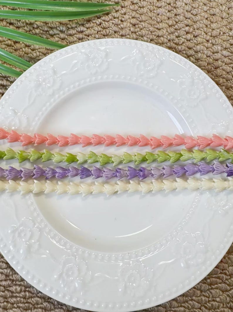 May include: Four strands of small, molded plastic flower beads arranged horizontally on a white ceramic plate with an embossed floral rim. The strands are colored in soft shades of peach, light green, lavender, and cream, each featuring a repeating petal design.