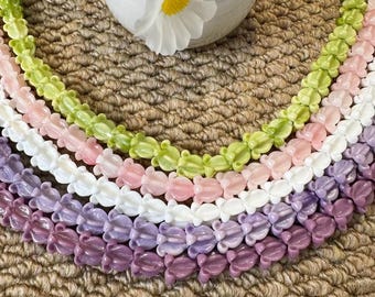 May include: A collection of colorful beaded necklaces in shades of green, pink, white, and purple. The beads are shaped like small flowers. The necklaces are arranged in a semi-circle on a textured, woven surface, with a white vase and white flowers in the background.