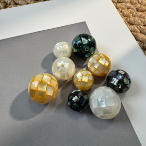 May include: A collection of spherical beads in various colors, including white, yellow, and dark green. The beads have a mosaic-like pattern, with each sphere composed of small, square-shaped pieces. The background is a mix of gray and white surfaces.