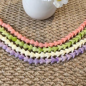 May include: Four strands of small, tulip-shaped beads arranged in a curved pattern. The strands are colored in soft pastel shades of peach, light green, cream, and lavender. Each bead features a delicate, multi-petaled floral design.
