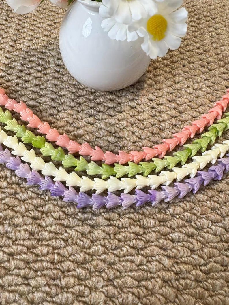 May include: Four strands of small, tulip-shaped beads arranged in a curved pattern. The strands are colored in soft pastel shades of peach, light green, cream, and lavender. Each bead features a delicate, multi-petaled floral design.