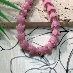 May include: A strand of light pink beads shaped like small flower buds arranged in a curved line. The word Lavender is printed in the upper left corner, and the text TimelessTreasuresHup is visible in the center of the bead arrangement.
