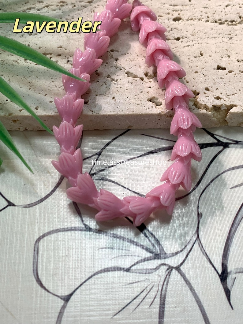 May include: A strand of light pink beads shaped like small flower buds arranged in a curved line. The word Lavender is printed in the upper left corner, and the text TimelessTreasuresHup is visible in the center of the bead arrangement.