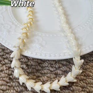 May include: A necklace composed of numerous off-white, flower-shaped beads strung together in a continuous strand. The beads feature a smooth, glossy finish and are arranged in a repeating pattern, creating a uniform, textured appearance throughout.