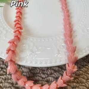 May include: A necklace composed of light pink beads, each carved into the shape of a small flower or tulip bud. The beads are strung together in a continuous strand, creating a uniform floral pattern throughout the length of the jewelry piece.