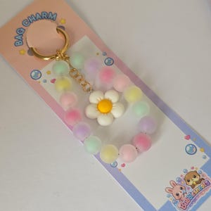 Könnte beinhalten: Ein pastellfarbener Taschenanhänger mit einem goldfarbenen Schlüsselring und einer Kette. Der Anhänger enthält ein Perlenarmband aus pastellfarbenen Kugeln und eine weiße Gänseblümchenblüte mit gelber Mitte. Die Verpackung trägt die Aufschrift "BAG CHARM" und "Bubblesnoots".