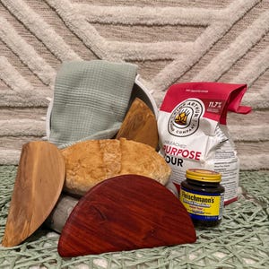 May include: A collection of baking supplies including a loaf of bread, wooden dough scrapers, a bag of flour, a jar of yeast, and a folded green towel. The flour bag reads "King Arthur Baking Company".