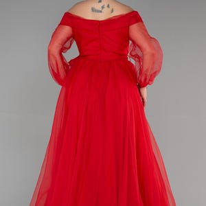 May include: A vibrant red, floor-length gown with an off-the-shoulder design and sheer, long sleeves. The dress has a full skirt and is made of a light, possibly chiffon-like material. The back of the dress is visible, showing the bodice's construction.