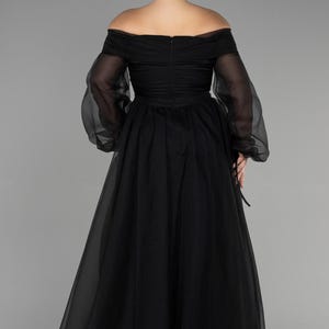 May include: Black off-the-shoulder formal gown with sheer long sleeves. The dress has a fitted bodice and a flowing skirt. The back of the dress has a zip closure. The model has blonde hair in a bun.