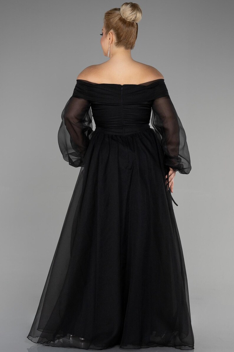 May include: Black off-the-shoulder formal gown with sheer long sleeves. The dress has a fitted bodice and a flowing skirt. The back of the dress has a zip closure. The model has blonde hair in a bun.